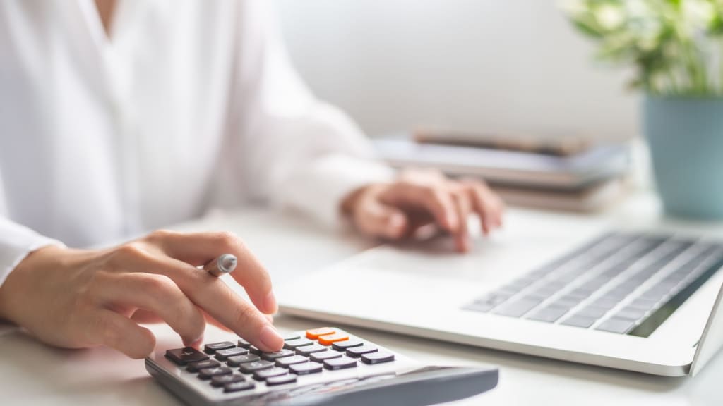 A person uses a calculator while working on a laptop at a desk, with books and a potted plant in the background.
